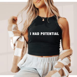 May include: Black tank top with the text "I HAD POTENTIAL" in white. The top is sleeveless and has a ribbed texture. The person is wearing a beige and white checkered cardigan.