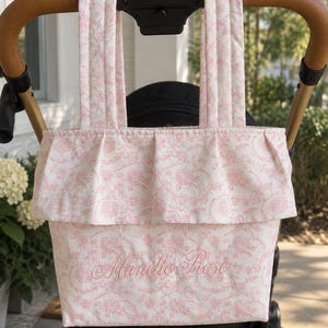 May include: A quilted fabric tote bag featuring a light pink floral pattern on a white background. The bag has two vertical straps, a ruffled top edge, and the name "Marielle Rose" embroidered in pink script on the front panel.