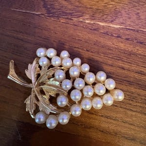 Op de afbeelding: Een goudkleurige broche in de vorm van een tros druiven met een bladaccent. De druiven worden gevormd door talloze kleine, ronde, witte kunstparels in een driehoekige opstelling, terwijl de bladeren van gestructureerd goudkleurig metaal zijn.