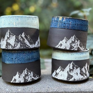 May include: Four ceramic mugs with a mountain design. The mugs are black with a white mountain range illustration. The rims are glazed in shades of blue and turquoise. The mugs are stacked on a stone surface.