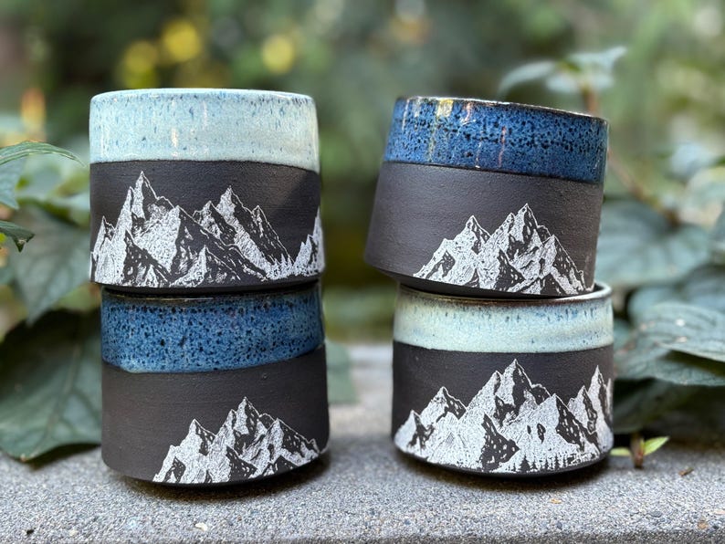 May include: Four ceramic mugs with a mountain design. The mugs are black with a white mountain range illustration. The rims are glazed in shades of blue and turquoise. The mugs are stacked on a stone surface.