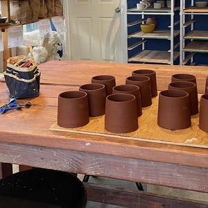 May include: A collection of unfinished, brown ceramic mugs arranged on a wooden board atop a large wooden table. The mugs are cylindrical with a slightly flared top. Pottery tools and supplies are visible in the background.