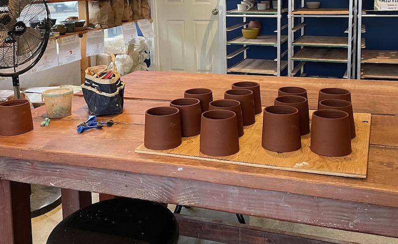 May include: A collection of unfinished, brown ceramic mugs arranged on a wooden board atop a large wooden table. The mugs are cylindrical with a slightly flared top. Pottery tools and supplies are visible in the background.