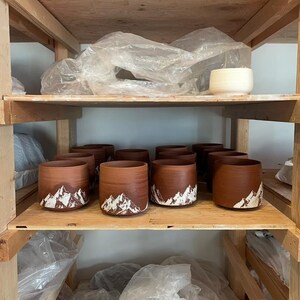 May include: A wooden shelf displays several handmade brown ceramic mugs with a mountain design in white. The mugs are cylindrical and have a rustic appearance. The shelf also holds other pottery and supplies.