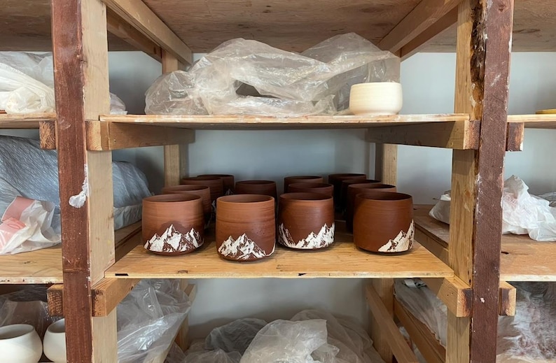 May include: A wooden shelf displays several handmade brown ceramic mugs with a mountain design in white. The mugs are cylindrical and have a rustic appearance. The shelf also holds other pottery and supplies.