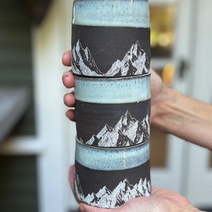 May include: A stack of four ceramic mugs. Each mug has a black band with a white mountain range design. The top and bottom rims have a light blue glaze. The mugs are held in a person's hands.