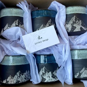 May include: A box of six ceramic mugs with a mountain design. The mugs have a black base with a white mountain graphic and a turquoise or blue rim. The mugs are nestled in white tissue paper, with a business card that reads "d. pottery design."