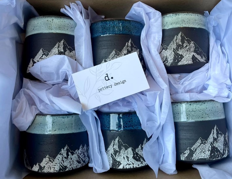 May include: A box of six ceramic mugs with a mountain design. The mugs have a black base with a white mountain graphic and a turquoise or blue rim. The mugs are nestled in white tissue paper, with a business card that reads "d. pottery design."