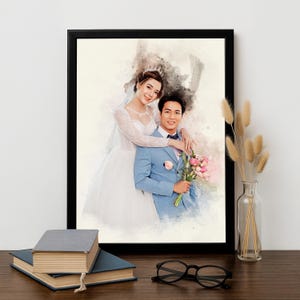 May include: A framed watercolour portrait of a newly married couple. The bride is in a white gown and veil, and the groom wears a blue suit. The artwork is in a black frame, with books and glasses in the foreground.