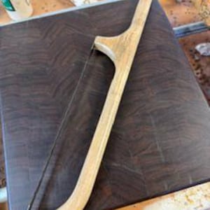 Könnte beinhalten: Eine Holzbügelsäge mit einem hellen, geschwungenen Griff und einem dünnen Metallblatt, die diagonal auf einem quadratischen, dunkelbraunen Holzschneidebrett mit sichtbarer Hirnholzmaserung und glatter, polierter Oberfläche liegt.