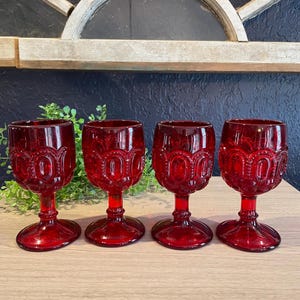 May include: Four ruby red goblets with ornate, embossed detailing. The stemmed glasses are arranged on a wooden surface, with a decorative window frame in the background. The glasses are a deep, transparent red.