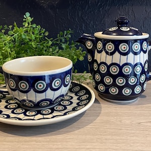 May include: A ceramic tea set featuring a blue and white pattern with a cup, saucer, and teapot. The design includes concentric circles in shades of blue, green, and red. The set is displayed on a light-colored surface.