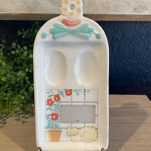 May include: A white ceramic spoon rest with a decorative floral and cat design. The top features a flower and bow design. The bottom has a cat and flower pot illustration. The spoon rest is supported by a metal stand.