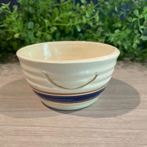 May include: Cream-colored ceramic yarn bowl with a blue and brown stripe design. The bowl has a curved shape with a slit for yarn. The bowl is sitting on a light-colored wooden surface.