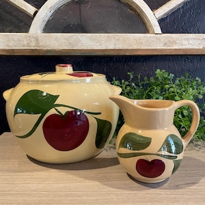May include: A cream-colored ceramic serving bowl with a lid and a small pitcher. Both pieces are decorated with a large red apple and green leaves design. The bowl has two handles and the pitcher has a handle and a spout.