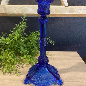 May include: A cobalt blue glass candlestick holder. The candlestick has a decorative base with a detailed floral design. The stem is a simple column with decorative rings. The top has a spiral design. The candlestick is approximately 10 inches tall.