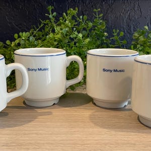 May include: Four white ceramic mugs with blue trim and the "Sony Music" logo. The mugs have a classic design with a handle and a sturdy base. They are arranged on a wooden surface with greenery in the background.