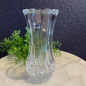May include: A clear glass vase with a flared top and a diamond-cut pattern on the lower portion. The vase has a scalloped rim and a curved, elegant shape. The vase is empty and sits on a white marble surface.