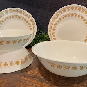 May include: A set of white dinnerware featuring a repeating golden-yellow floral pattern. The collection includes two round plates and two deep bowls, with one bowl inverted to show the exterior design and the other bowl upright to show the interior.