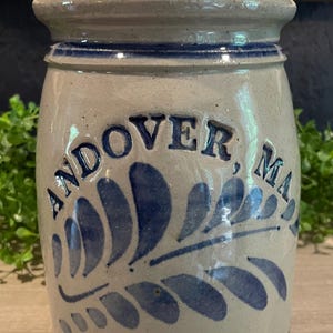 May include: A cylindrical stoneware crock with a light gray speckled finish and a flared rim. The front features cobalt blue hand-painted leaf motifs and the text "ANDOVER, MA." above the text "EST. 1646" in a bold, dark blue serif font.