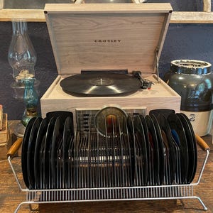 May include: A wooden Crosley record player with a black vinyl record on the turntable. A chrome record holder with wooden handles holds several black vinyl records. A vintage oil lamp and ceramic jar are in the background.