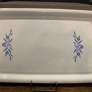May include: A rectangular white ceramic serving tray featuring a glossy finish and a raised border. The center of the tray is decorated with two symmetrical blue floral patterns, each consisting of small flowers and leafy stems on opposite sides.