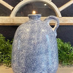 May include: A blue and white speckled ceramic jug with a handle. The jug has a small opening at the top and a lit candle inside. The background includes a wooden window frame and greenery.