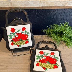 May include: Two black cast iron trivets shaped like teapots. Each trivet has a white tile insert with a colorful illustration of red apples, tulips, and berries in a yellow basket. The trivets are on a light wood surface.