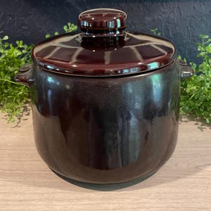 May include: A dark brown ceramic casserole dish with a matching lid. The dish has a rounded shape with small handles on either side. The lid has a knob on top for easy handling. The casserole dish is likely used for cooking and serving food.