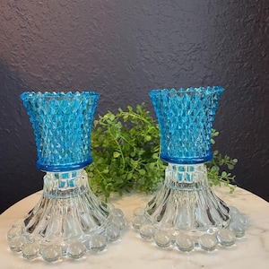 May include: Two vintage glass candle holders. The top portion is a vibrant turquoise blue with a diamond pattern. The base is clear glass with a beaded design around the bottom. The candle holders are displayed on a white marble surface.
