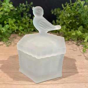 May include: A frosted glass trinket box with a bird figurine on top. The box is square with a textured basket weave design. The bird is perched on the lid, facing left. The background features greenery.