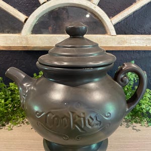 May include: A dark brown ceramic cookie jar in the shape of a teapot. The word "Cookies" is embossed on the front. The jar has a lid and a handle. The jar is sitting on a wooden surface.