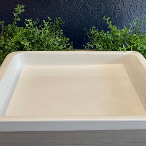 May include: A rectangular, off-white ceramic baking dish with slightly raised sides. The dish is empty and sits on a wooden surface, with green foliage in the background. Ideal for baking casseroles or roasting vegetables.