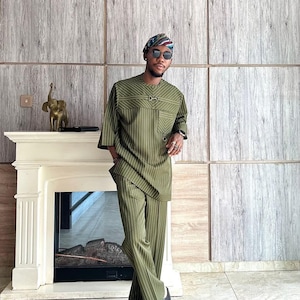 May include: Olive green pinstripe suit, consisting of a short-sleeved tunic and matching trousers. The outfit is paired with black sandals and a patterned head wrap. The background features a fireplace and a wooden panel wall.
