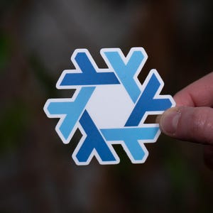 May include: A sticker with a geometric design in shades of blue, resembling a snowflake or a stylized six-pointed star. The design is composed of interlocking shapes with a white background and a white border.