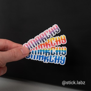 Op de afbeelding: Drie kleurrijke stickers met het woord "ANARCHY" in een pixelachtige lettertype. De stickers zijn in tinten paars, oranje en blauw. De stickers worden tegen een zwarte achtergrond gehouden.