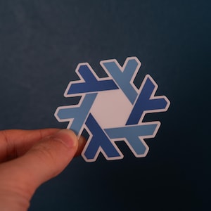 Könnte beinhalten: Ein Aufkleber mit einem Schneeflocken-Design. Die Schneeflocke besteht aus ineinandergreifenden Formen in Blau- und Weißtönen mit einem weißen Zentrum. Der Aufkleber wird vor einem dunkelblauen Hintergrund gehalten.