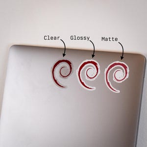 Puede incluir: Tres pegatinas con un dise&ntilde;o de espiral rojo y blanco se muestran en un port&aacute;til. Las pegatinas est&aacute;n etiquetadas como "Clear", "Glossy" y "Matte" para mostrar los diferentes acabados. Las pegatinas son un art&iacute;culo decorativo.