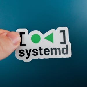 Puede incluir: Una pegatina blanca con la palabra "systemd" en negro, con un diseño de círculo y triángulo verde. La pegatina se sostiene sobre un fondo verde azulado.