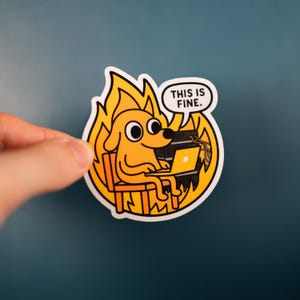 May include: A sticker depicting a cartoon dog in a chair, engulfed in flames, working on a laptop. A speech bubble above the dog reads, "THIS IS FINE." The sticker's colours are yellow, orange, black, and white.