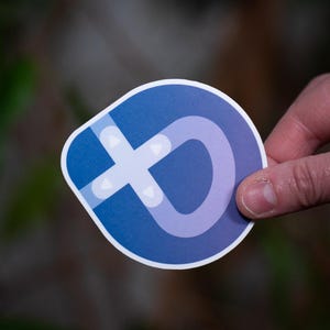 Op de afbeelding: Een sticker met een afgeronde vorm, met een blauw en paars ontwerp. Het ontwerp bevat een witte kruisvorm en een gebogen lijn. De sticker wordt door een hand tegen een vage achtergrond gehouden.