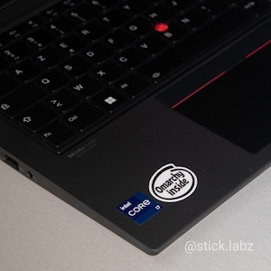 Può includere: Laptop nero con tastiera e touchpad. Il laptop ha un trackpoint rosso e due adesivi: uno con il logo Intel Core i7 e l'altro con la scritta "Omarchy Inside". Il laptop è su una superficie chiara.