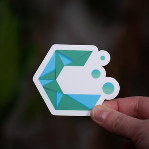 Peut inclure: Un autocollant blanc avec un motif géométrique bleu et vert. L'autocollant a une forme hexagonale avec des triangles superposés et trois petits cercles. L'autocollant est tenu par une main sur un fond flou.