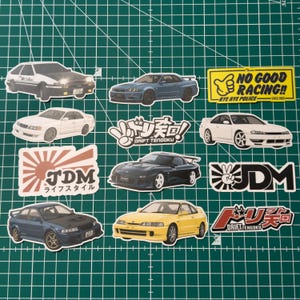 Peut inclure: Collection d'autocollants de voitures JDM (Japanese Domestic Market). Les autocollants présentent divers modèles de voitures de différentes couleurs, ainsi que du texte tel que "JDM", "DRIFT TENGOKU" et "NO GOOD RACING!! BYE BYE POLICE SINCE 1985".