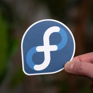 Peut inclure: Un autocollant avec un fond bleu foncé et une lettre « f » stylisée blanche et bleue. L'autocollant, de forme rectangulaire arrondie, est tenu par une main.