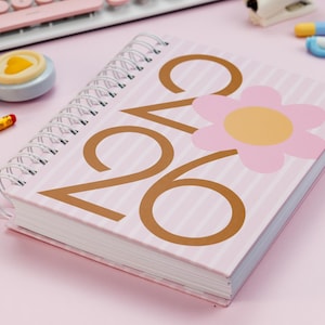 Puede incluir: Un cuaderno de espiral a rayas rosas y blancas con los números "26" en marrón y un gráfico de flor rosa. El cuaderno está rodeado de material de oficina, incluyendo un teclado, cinta adhesiva y bolígrafos. El cuaderno está diseñado para la planificación y organización.