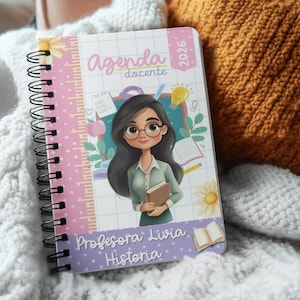 Puede incluir: Una agenda con espiral con un diseño floral rosa y blanco. La portada presenta una ilustración de una mujer con gafas sosteniendo un libro, con el texto "Agenda docente 2026" y "Professora Livia Historia".