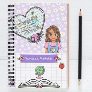 Pode incluir: Caderno espiral com um design floral e quadriculado. A capa apresenta uma ilustração de desenho animado de uma professora e o texto "El quería cambiar el mundo y por eso se convirtió en maestra." Um lápis preto repousa ao lado do caderno.