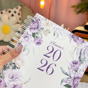 Pode incluir: Um planejador floral para o ano de 2026. A capa apresenta um fundo branco com rosas roxas e folhas verdes. A palavra "Planner" e os números "20 26" são impressos em roxo. O planejador é segurado por uma mão.