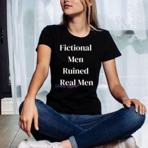 May include: Black t-shirt with the text "Fictional Men Ruined Real Men" in white. The shirt is worn with blue jeans. The model is sitting on a wooden floor.